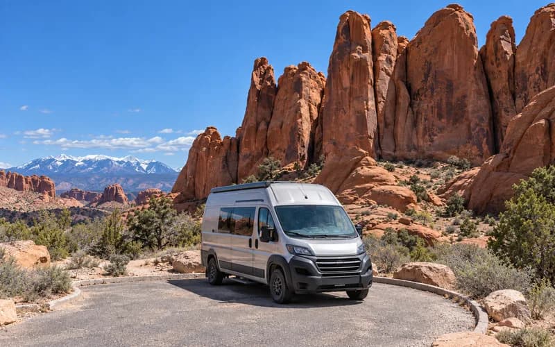 A camper van in a slickrock parking area with sandstone fins rising behind it