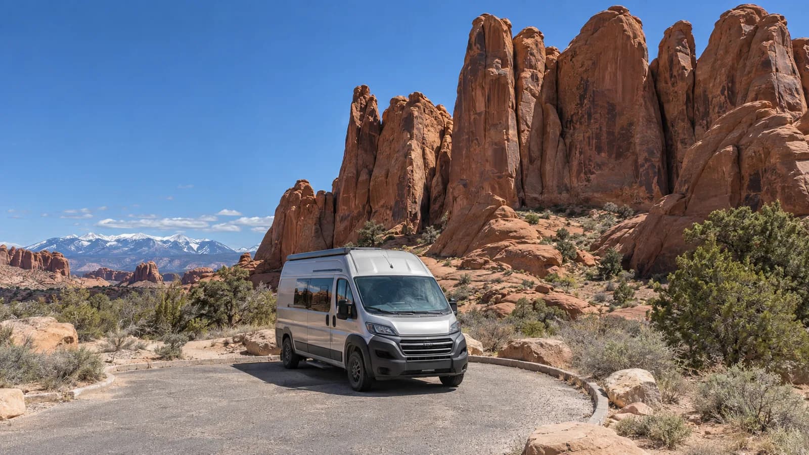 A camper van in a slickrock parking area with sandstone fins rising behind it